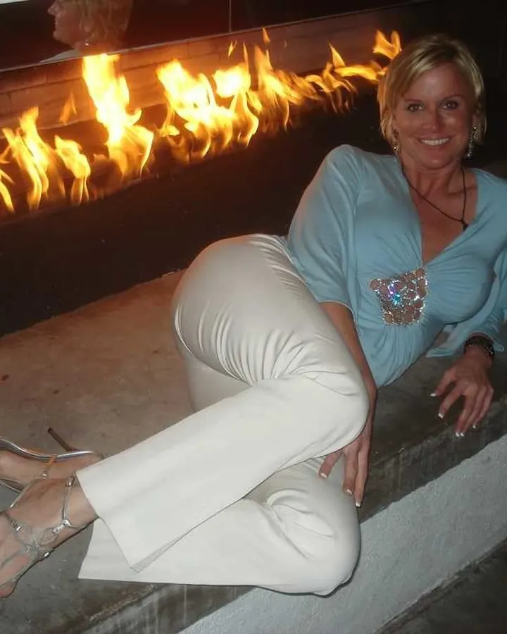 Femme blonde en tenue bleue et pantalon blanc, assise sur un rebord devant un feu de cheminée, souriante, les jambes croisées, chaussures à talons argentées.