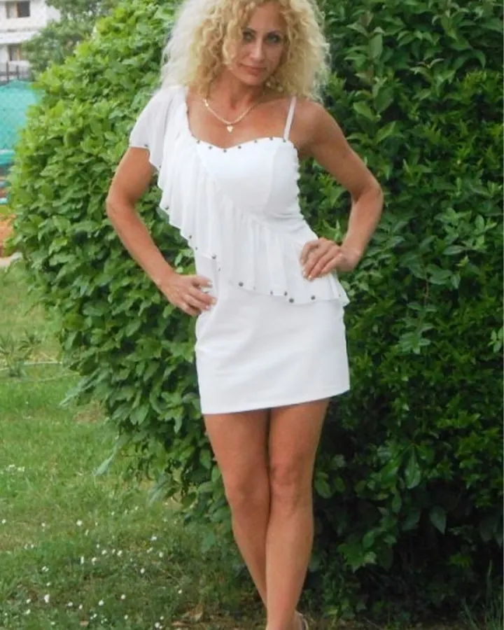 Femme blonde en robe blanche courte, debout devant un buisson vert, mains sur les hanches