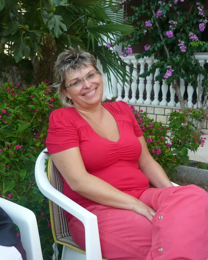 Femme souriante en tenue rose, assise sur une chaise blanche devant des plantes fleuries