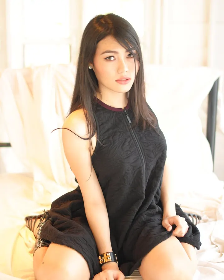 Femme asiatique en robe noire, assise sur un lit blanc, regardant l'appareil