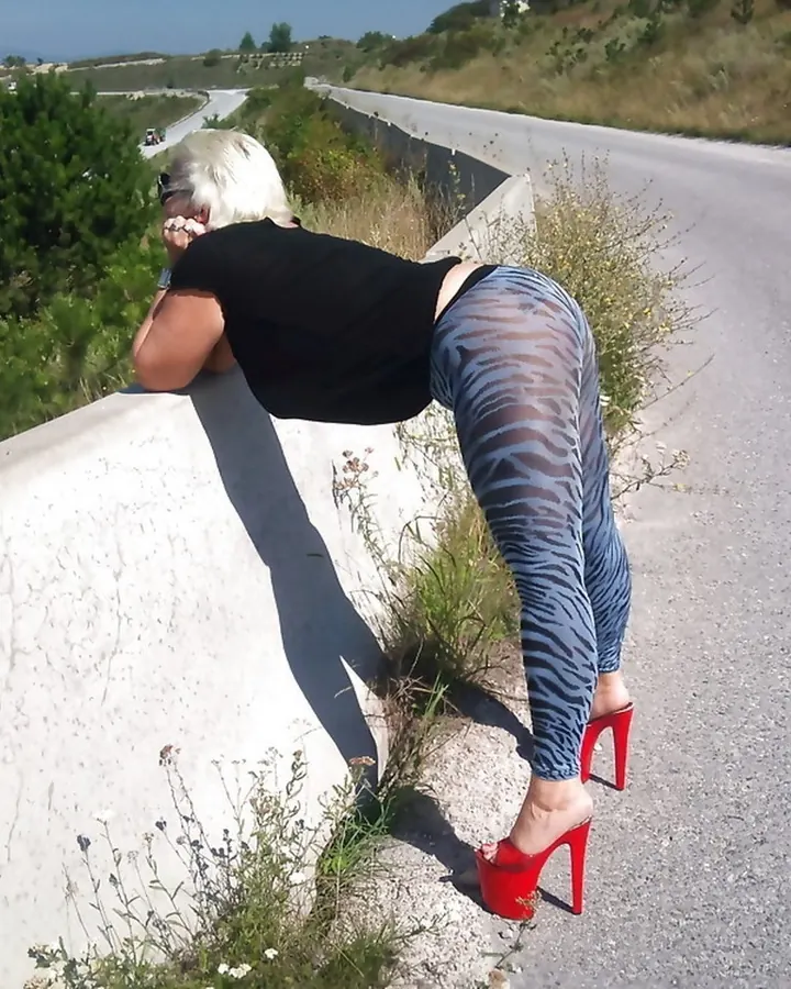 Femme blonde vue de dos, portant un haut noir et des leggings zébrés, chaussée de talons rouges, appuyée sur un mur près d’une route.
