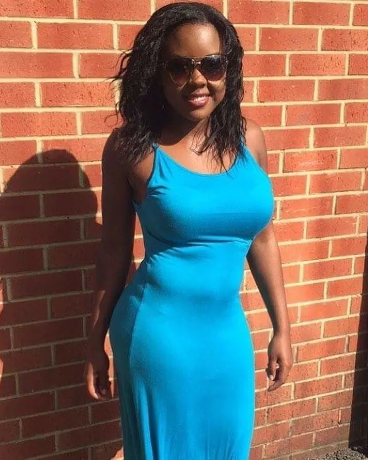 Femme noire souriante, portant une robe bleue, debout devant un mur de briques.