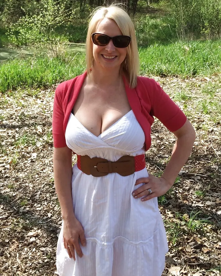 Femme blonde souriante, portant une robe blanche avec ceinture marron, debout dans un jardin ensoleillé.