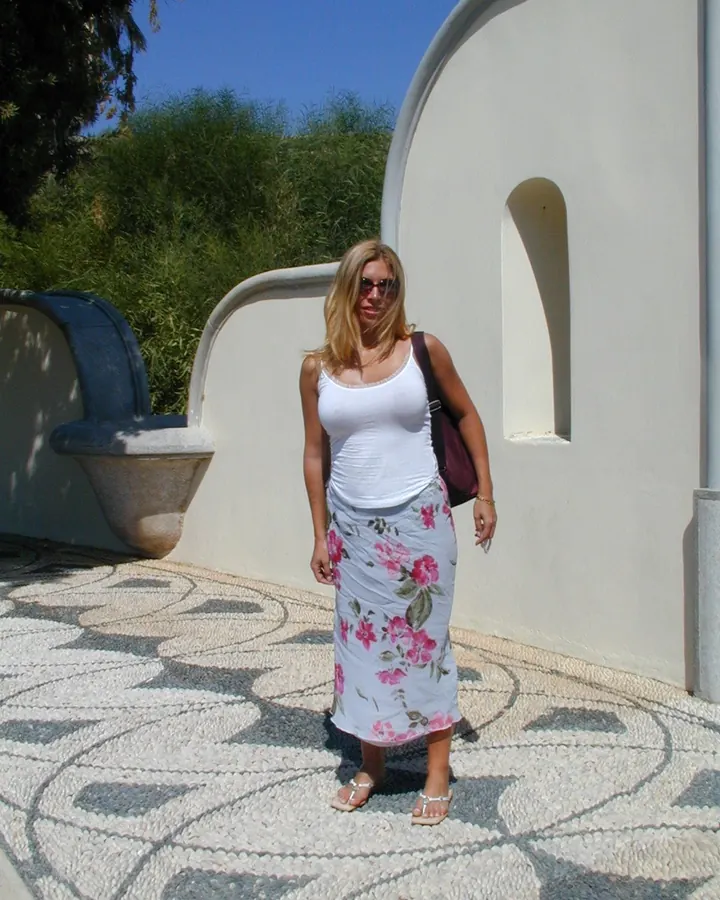 Femme blonde souriante, portant une robe blanche avec ceinture marron, debout dans un jardin ensoleillé.