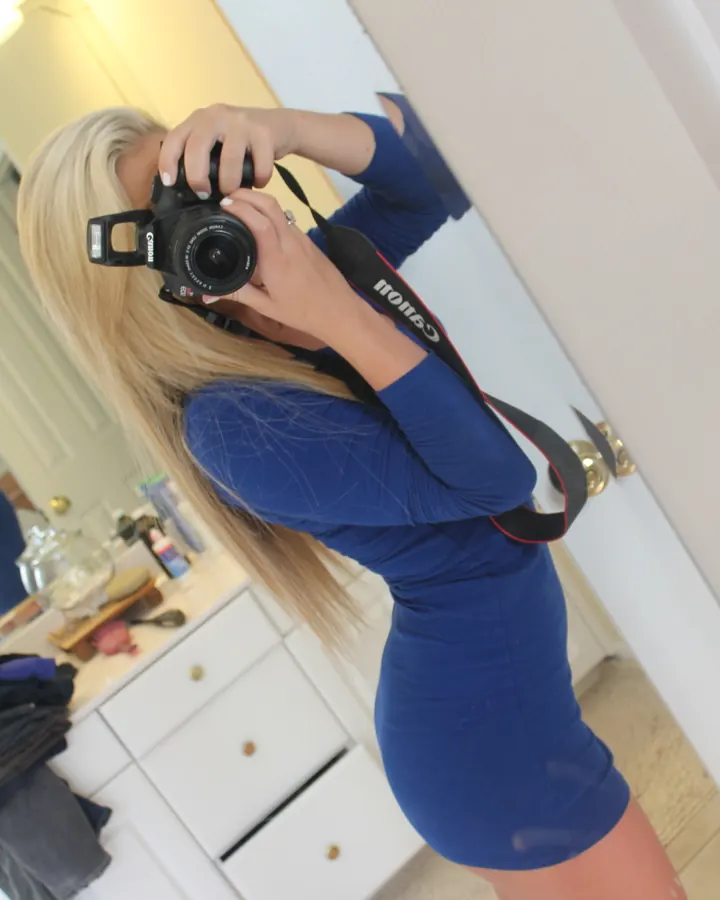 Femme blonde portant une robe bleue, prenant un selfie devant un miroir dans une salle de bain, tenant un appareil photo Canon.