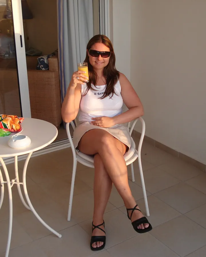 Femme aux cheveux bruns portant un haut blanc et une jupe beige, assise à une table blanche sur une terrasse, tenant un verre de jus d'orange.