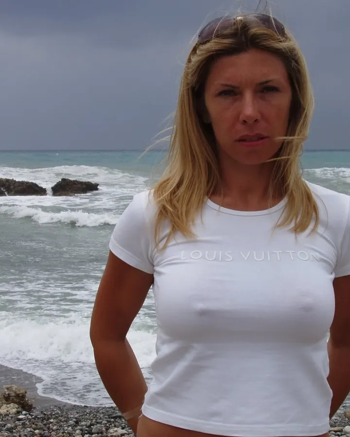 Femme blonde en t-shirt blanc Louis Vuitton sur une plage rocheuse, regardant l'appareil photo avec un air sérieux.