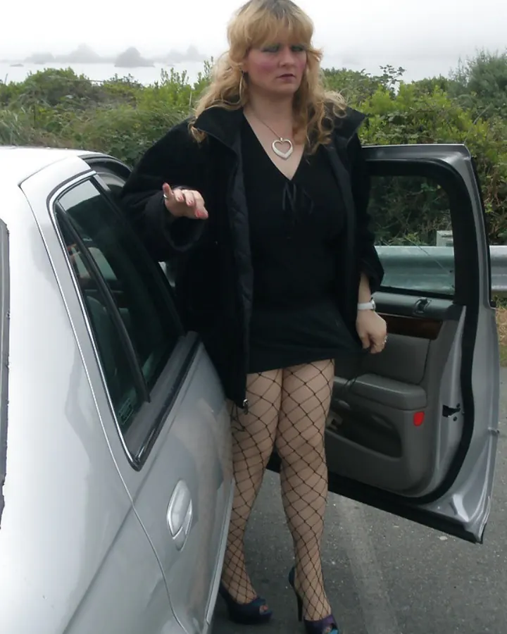 Femme aux cheveux blonds portant un t-shirt blanc et une jupe beige, assise à une table blanche sur une terrasse, tenant un verre de jus d'orange.