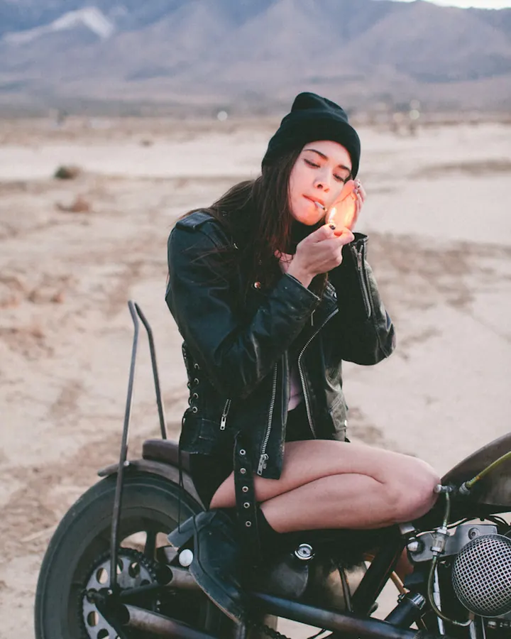 Femme en veste en cuir noire et bonnet, assise sur une moto dans un désert.