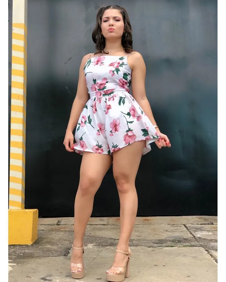 Jeune femme brune aux cheveux châtains, portant une robe fleurie blanche et rose, debout contre un mur noir, les mains sur les hanches.