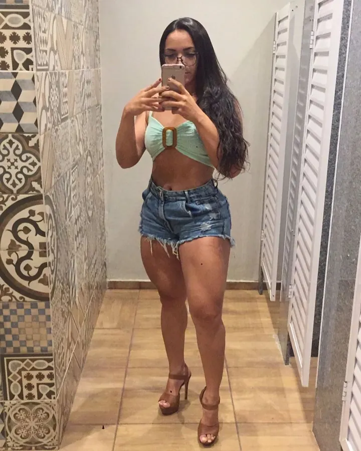 Femme aux cheveux longs noirs, portant un haut vert menthe avec une boucle marron, un short en jean déchiré et des sandales compensées, prenant un selfie devant un mur carrelé.