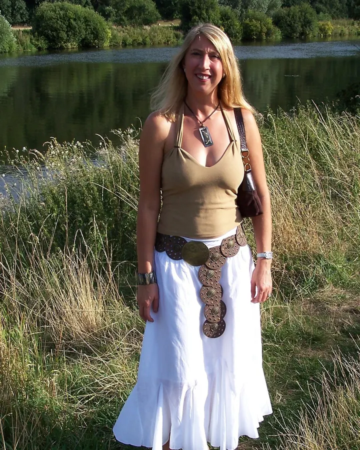Femme blonde souriante, portant un haut beige et une longue jupe blanche, debout près d'un lac, avec un sac à main en bandoulière.
