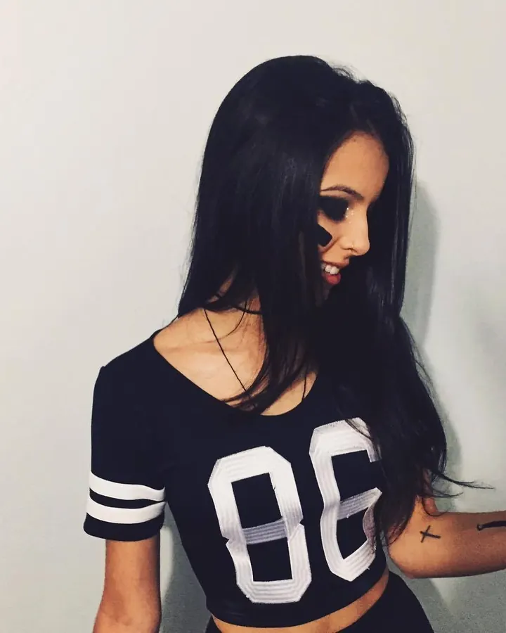 Femme latina aux cheveux bruns, en lingerie noire, posant sur un lit avec un regard intense.