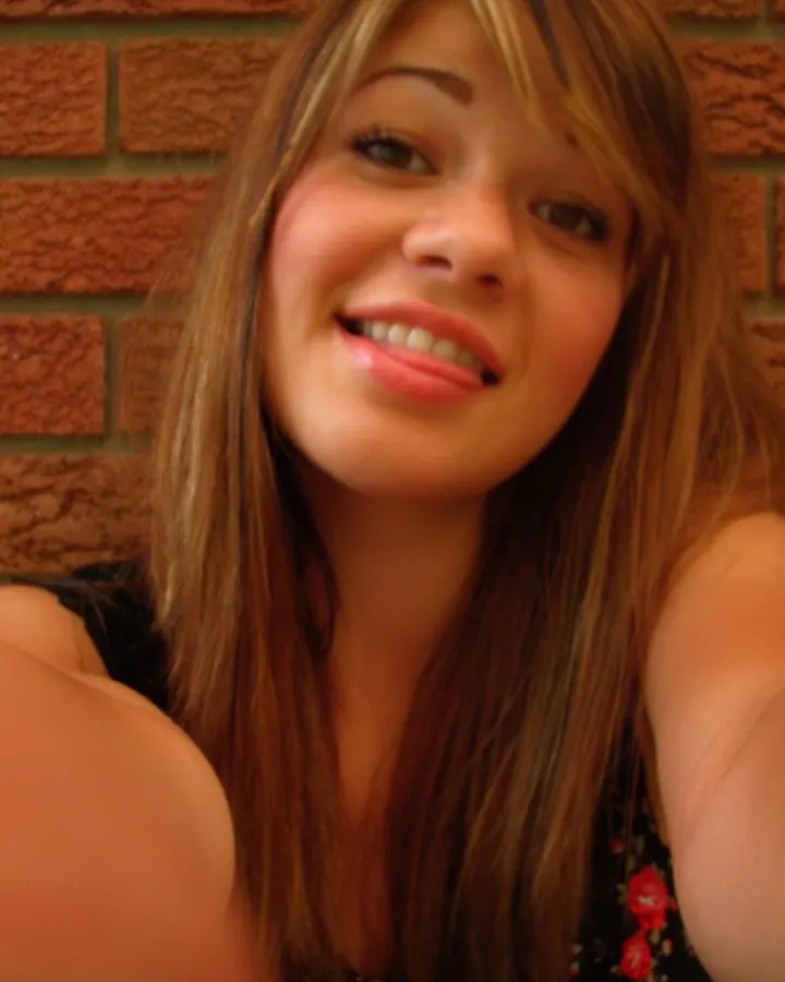 Jeune femme aux cheveux bruns, souriante, en train de prendre un selfie devant un mur en briques rouges.