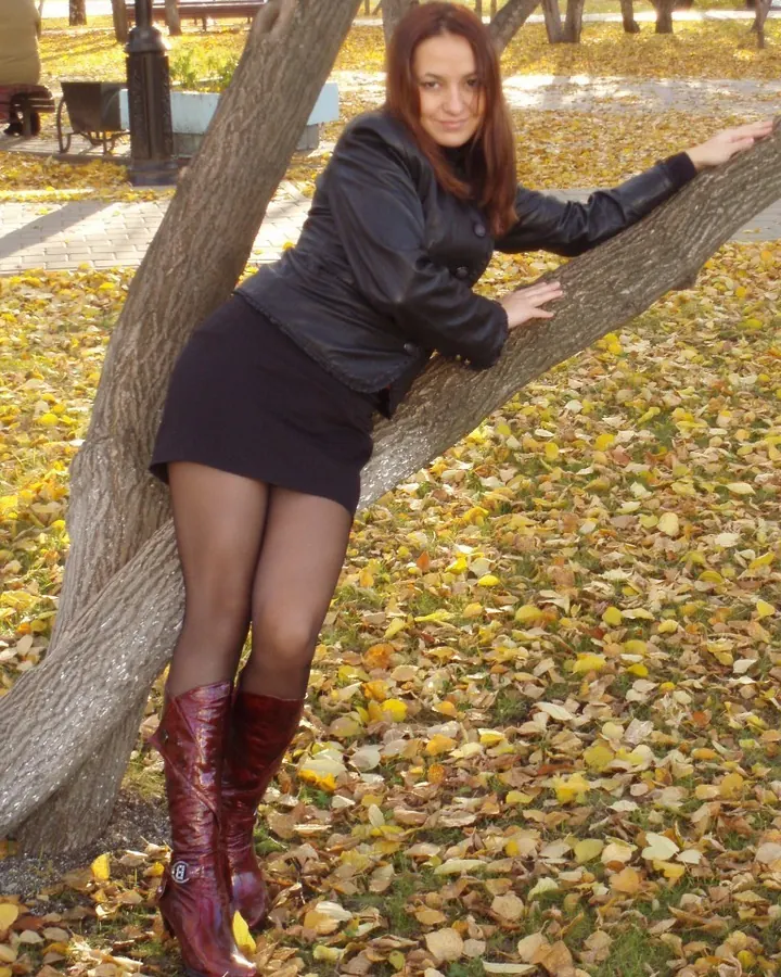 Femme aux cheveux roux, veste noire, jupe courte, bottes rouges, appuyée sur un arbre en automne.