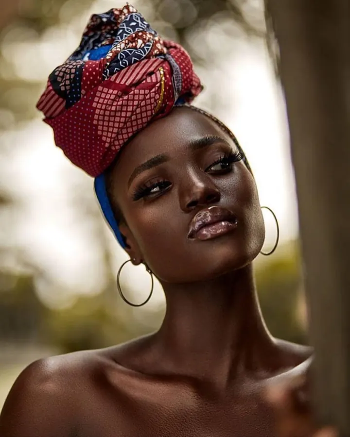 Femme noire avec un turban coloré, regard intense, boucles d'oreilles fines, peau luisante.