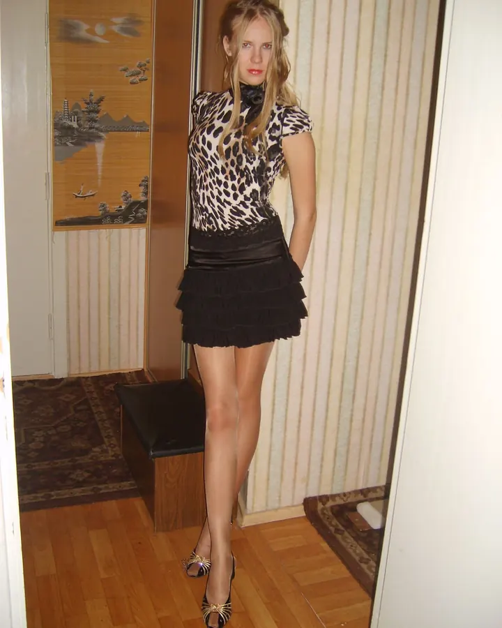 Jeune femme blonde, en robe noire à imprimé léopard, jupe noire frangée, chaussures à talons, debout dans un couloir, regardant l'appareil.