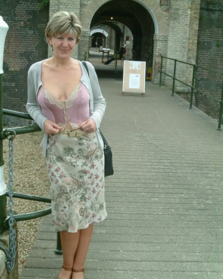 Femme aux cheveux blonds courts en haut rose, cardigan gris, jupe fleurie, debout sur un pont en pierre.