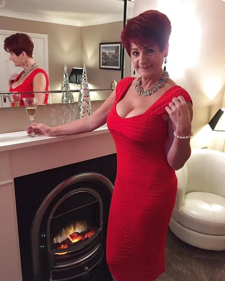 Femme aux cheveux roux courts en robe rouge, tenant un verre de vin, devant une cheminée allumée.