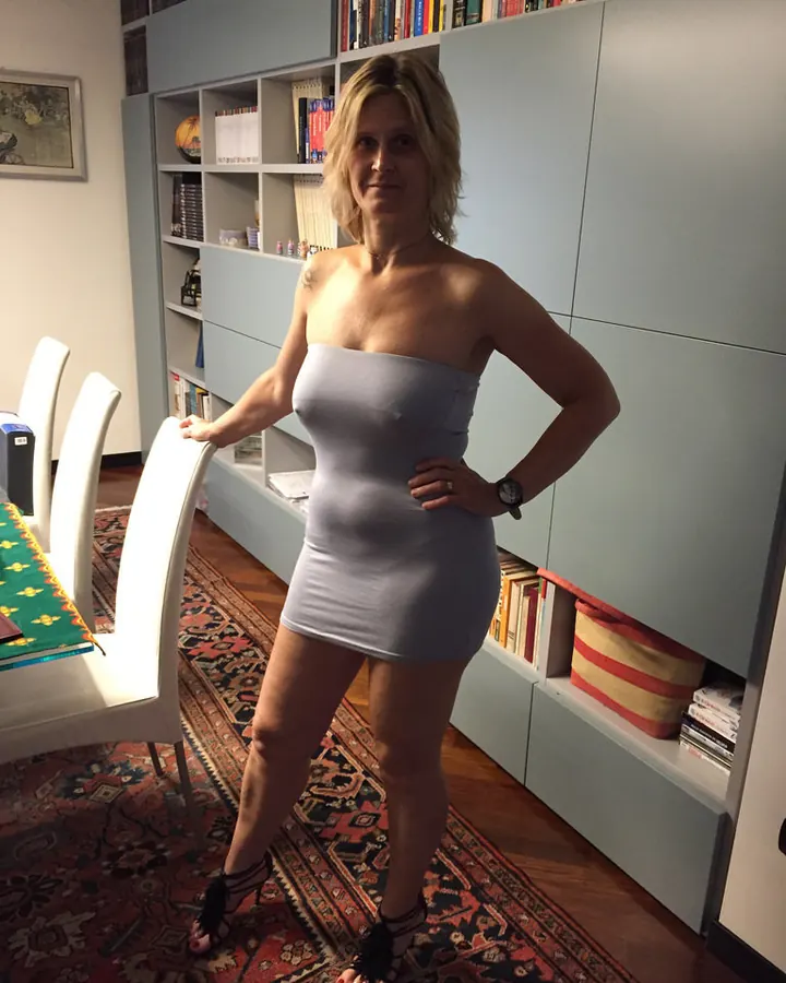 Femme aux cheveux platine portant une robe grise ajustée, debout dans un salon avec une bibliothèque en arrière-plan.