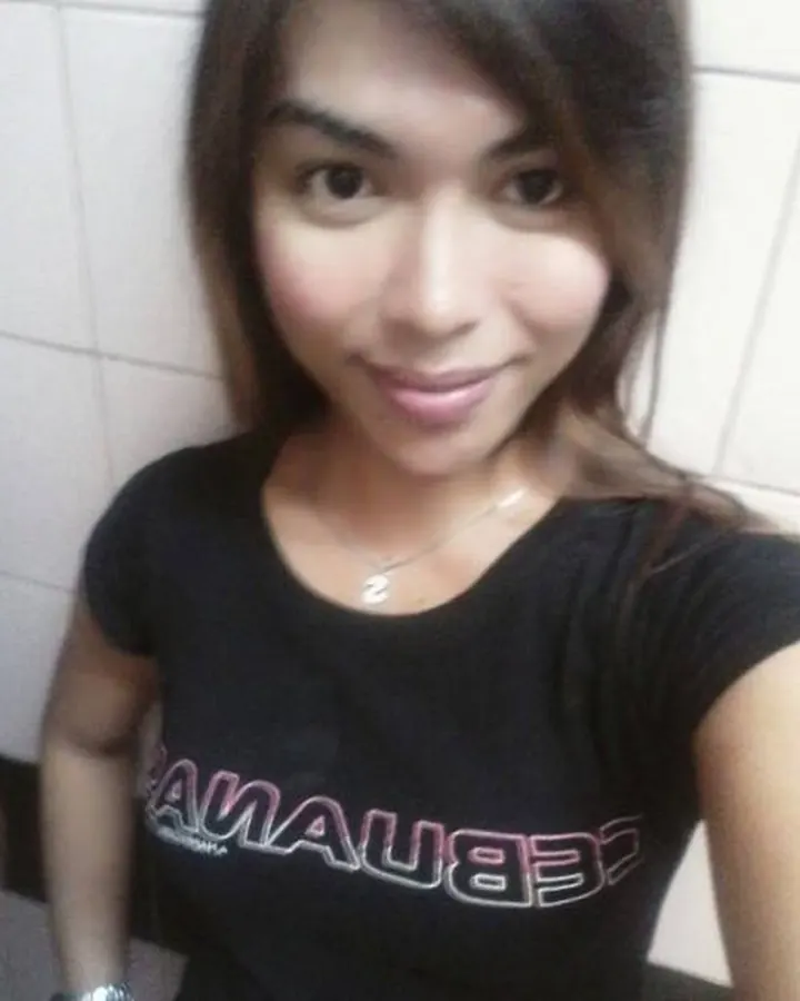 Femme aux cheveux bruns foncés, souriant à la caméra, portant un t-shirt noir avec un logo inversé, assise contre un mur carrelé blanc.