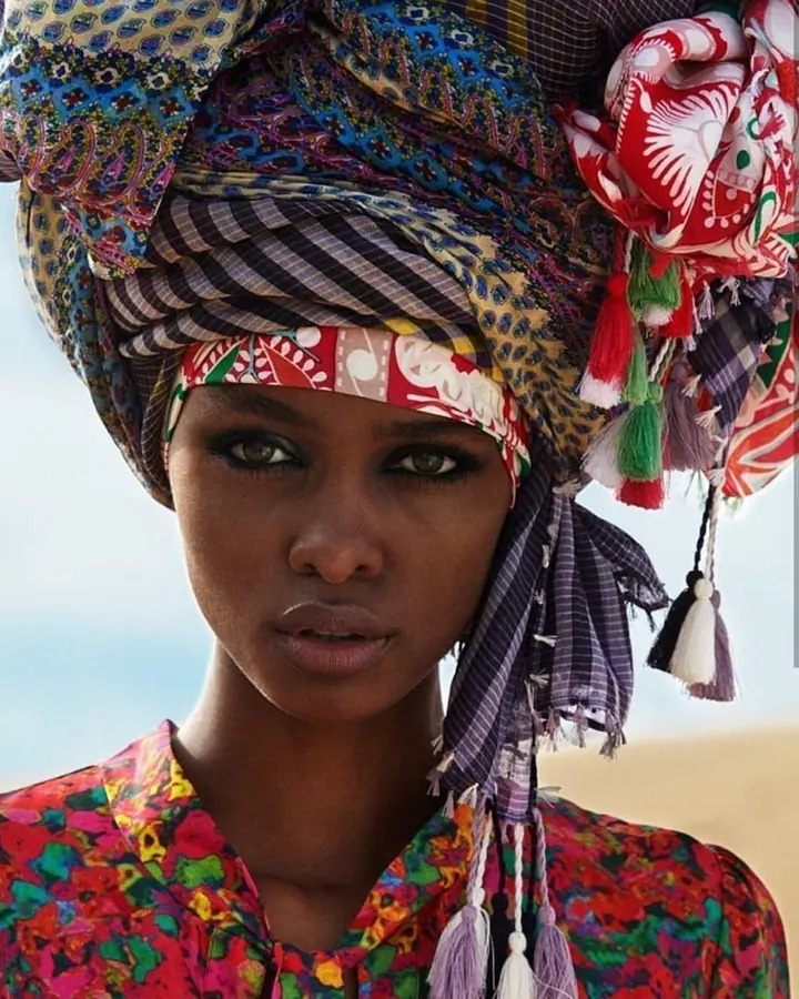 Femme noire avec un turban coloré et un haut multicolore, regardant directement la caméra avec un air sérieux, sur fond flou de plage.