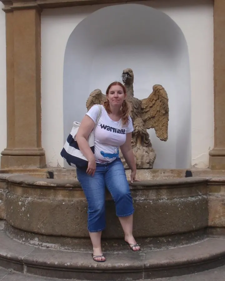 Femme blonde aux cheveux longs, portant une tenue rose vif moulante avec une ceinture métallique, se tenant près d'une étagère blanche dans une pièce aux murs bleus.