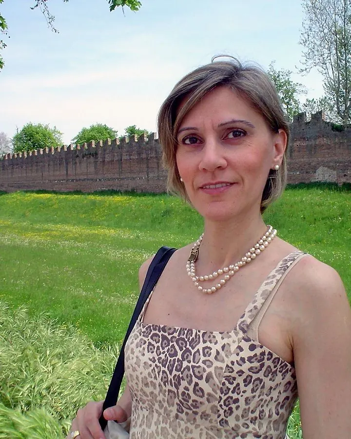 Femme aux cheveux gris courts, souriant à la caméra, portant une robe léopard et un collier de perles, debout dans un parc verdoyant avec un mur de briques en arrière-plan.