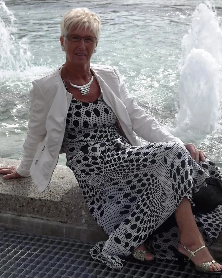 Femme aux cheveux blancs assise près d'une fontaine, portant une robe à pois noirs et blancs