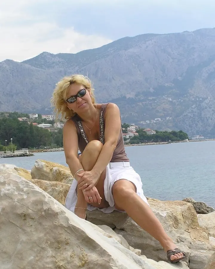Femme blonde aux cheveux courts, assise sur des rochers près de la mer, lunettes de soleil, sourire espiègle