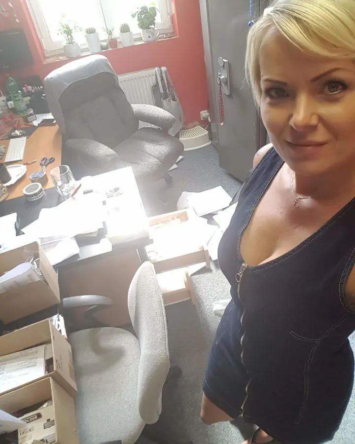 Femme blonde en robe bleue, debout dans un bureau encombré, regardant l'appareil, souriante