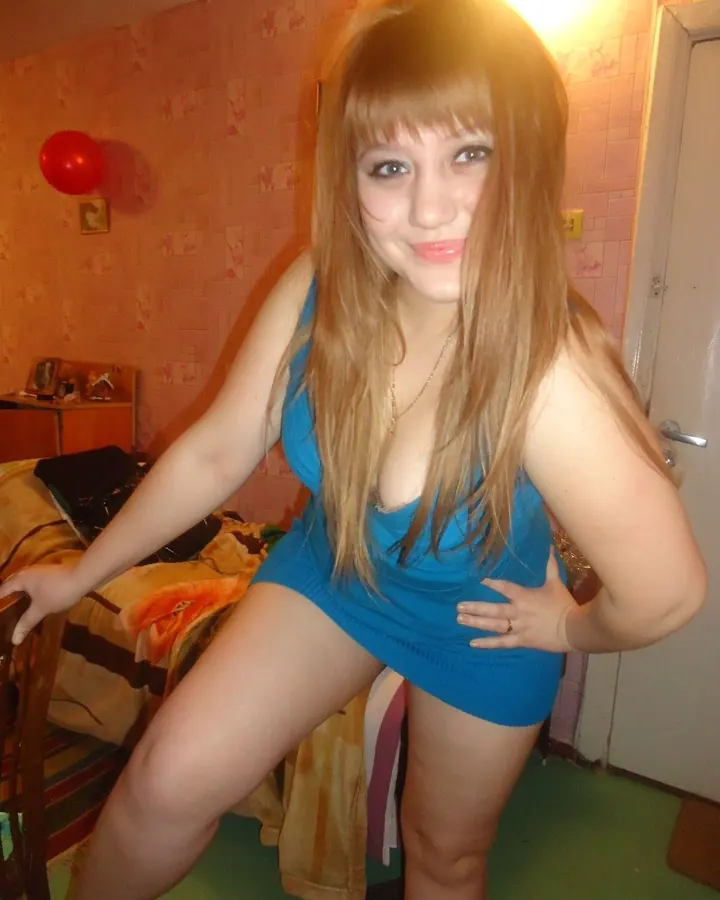 Femme blonde en robe bleue courte, souriante, pose dans une chambre aux murs roses.