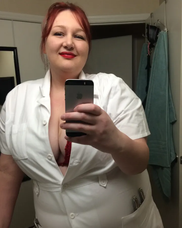 Femme rousse en tenue blanche de nurse, prenant un selfie dans un miroir, soutien-gorge rouge visible.