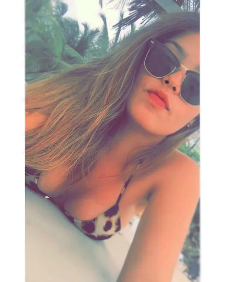 Femme blonde en haut noir, posant de dos sur un lit, mains dans les cheveux, culotte noire à rayures blanches.