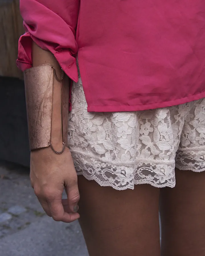 Femme portant un short court en dentelle blanche sous une chemise rose, avec un bracelet métallique au poignet, vue de dos.