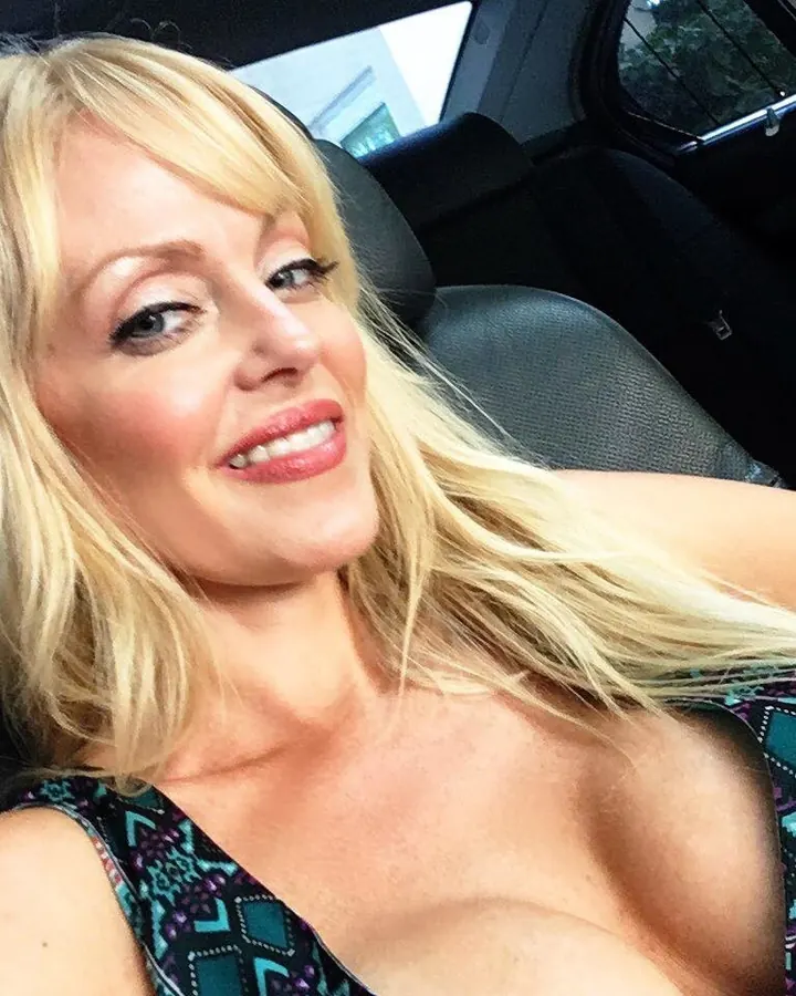 Femme blonde aux yeux bleus, souriante, assise dans une voiture, portant un haut coloré, regardant l'objectif.