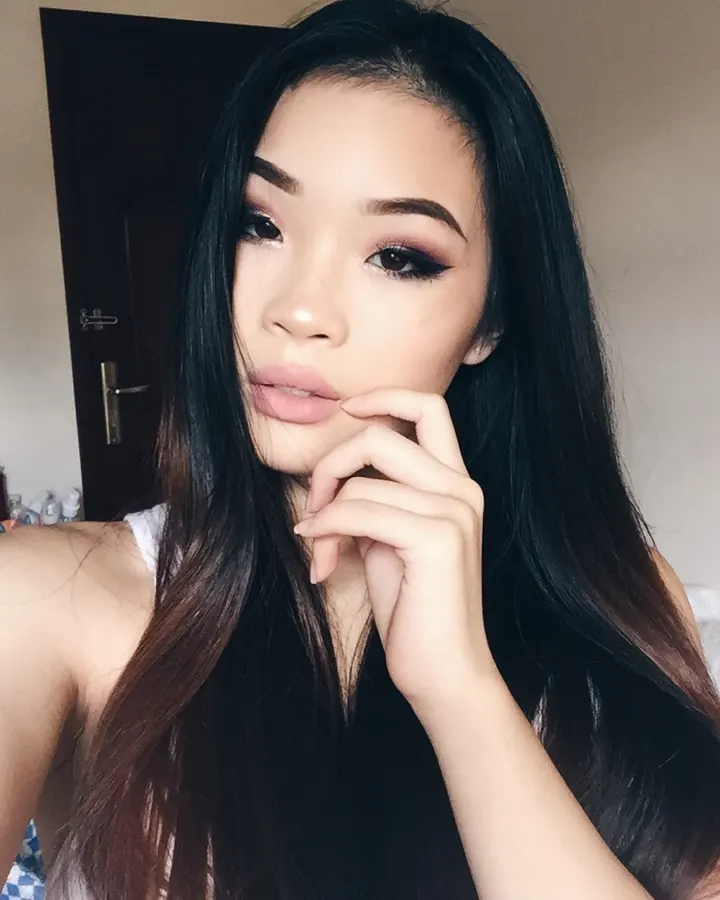 Jeune femme aux cheveux noirs longs, maquillage prononcé, regardant l'objectif, la main près de la bouche, dans une pièce intérieure.