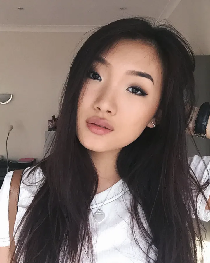 Jeune femme aux cheveux noirs longs, regardant l'objectif, portant un haut blanc, dans une pièce intérieure.