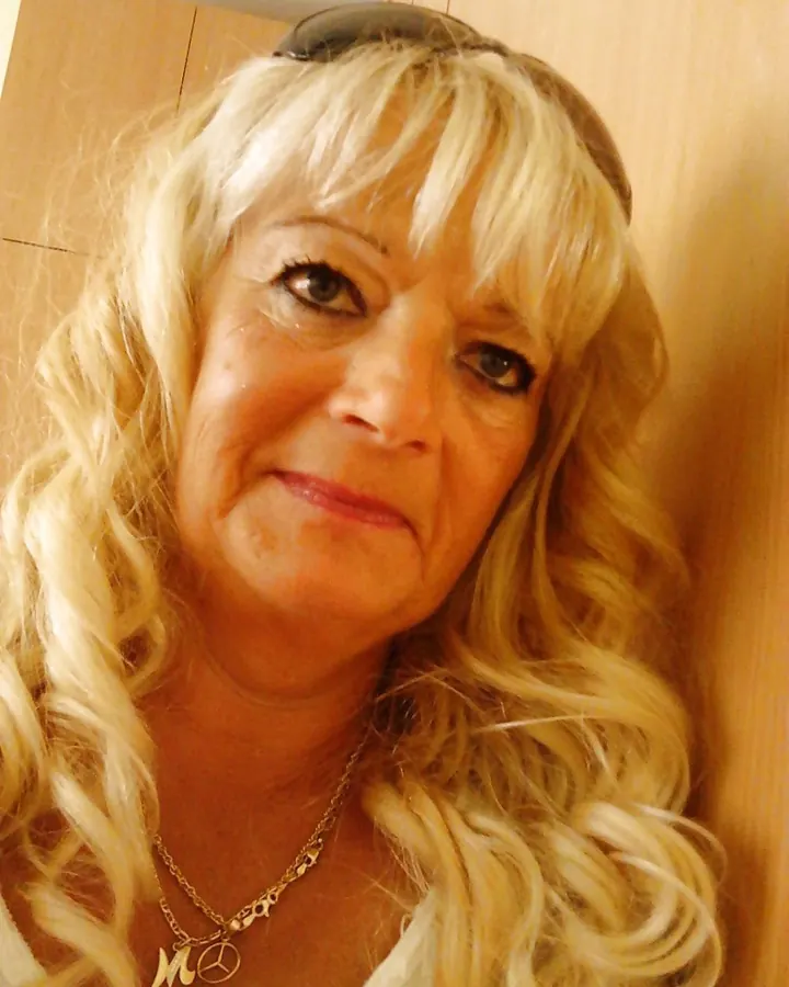 Femme âgée aux cheveux blonds platine, souriante, en gros plan, portant un collier avec un pendentif, devant un fond bois clair.