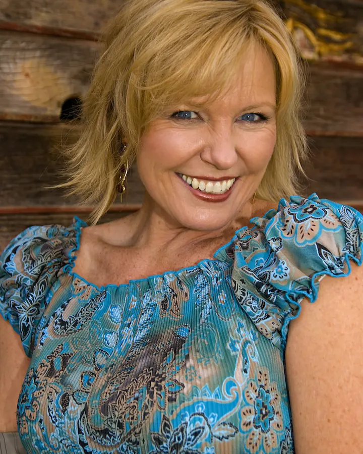 Femme aux cheveux blonds longs, souriante, en gros plan, portant un haut noir, dans une pièce intérieure.