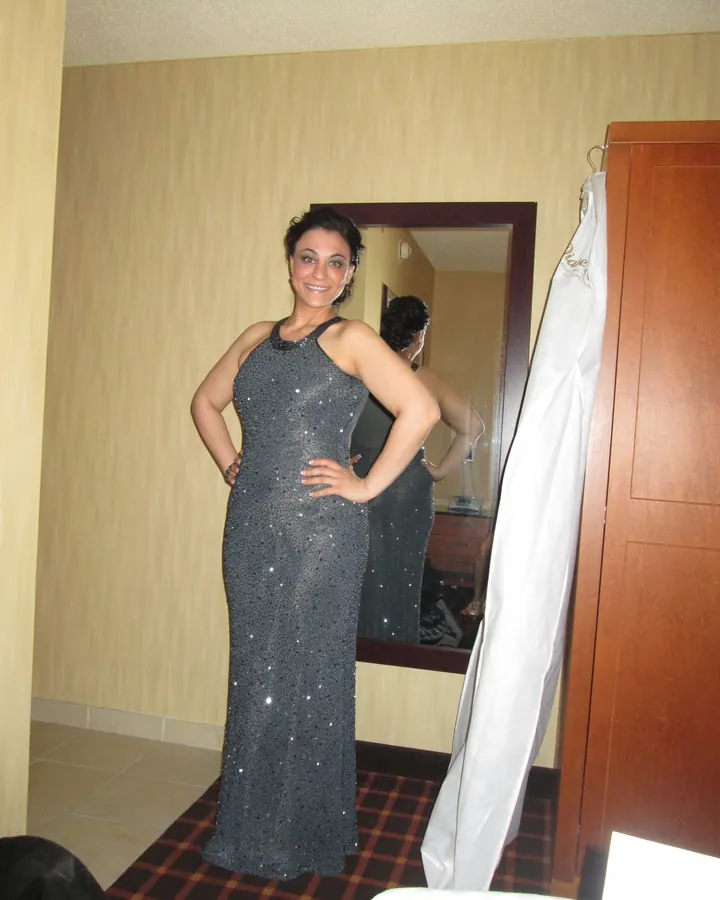 Femme aux cheveux noirs courts, souriante, debout devant un miroir dans une chambre, portant une robe longue argentée scintillante.