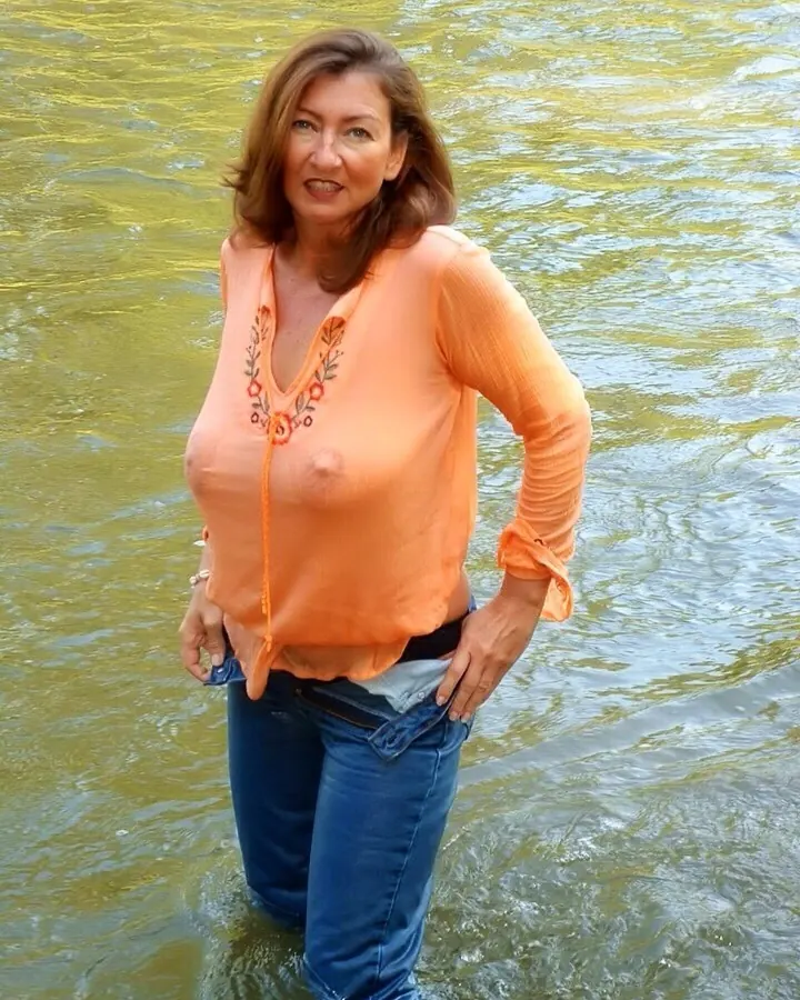 Femme aux cheveux bruns mi-longs, souriante, debout dans une pièce, portant un haut noir transparent et une jupe noire, devant un canapé blanc.