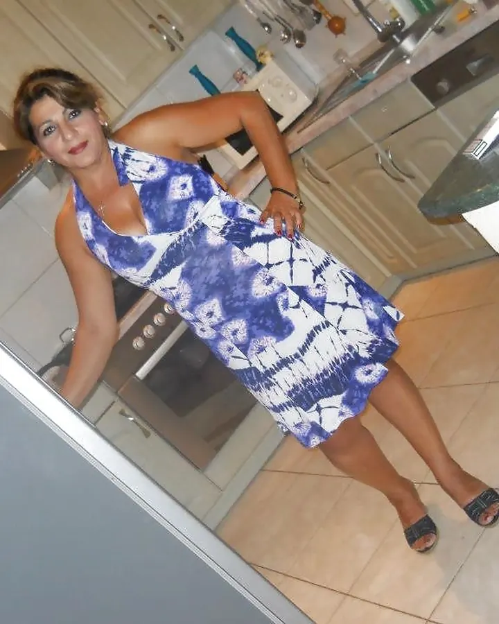 Femme aux cheveux bruns portant une robe bleue et blanche, posant dans une cuisine avec un four et une table en verre.