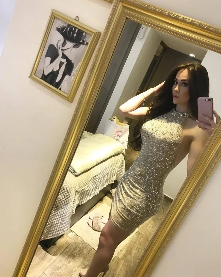 Femme aux cheveux bruns-portant un haut noir et un short blanc, prenant un selfie dans un miroir, avec un carrelage beige en arrière-plan.