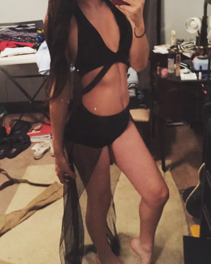 Jeune femme aux cheveux longs bruns en tenue noire, prenant un selfie dans une chambre encombrée, tenant un morceau de tissu transparent