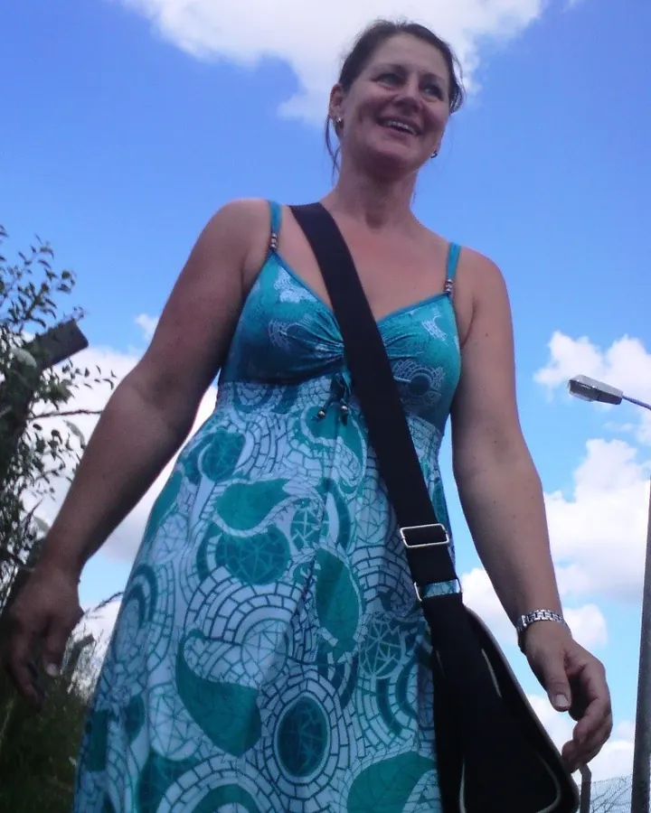 Femme souriante en robe turquoise, debout en extérieur sous un ciel bleu, portant un sac à bandoulière.