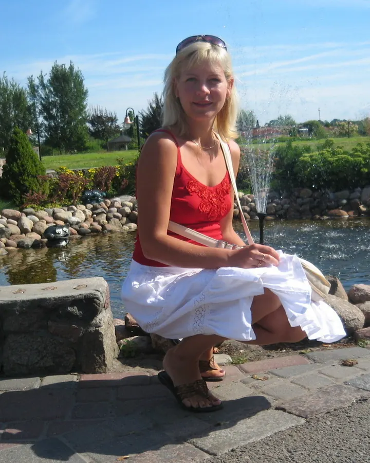 Femme aux cheveux blonds portant un haut noir transparent et un short en jean, posant sur un banc en pierre avec un fond de briques rouges.