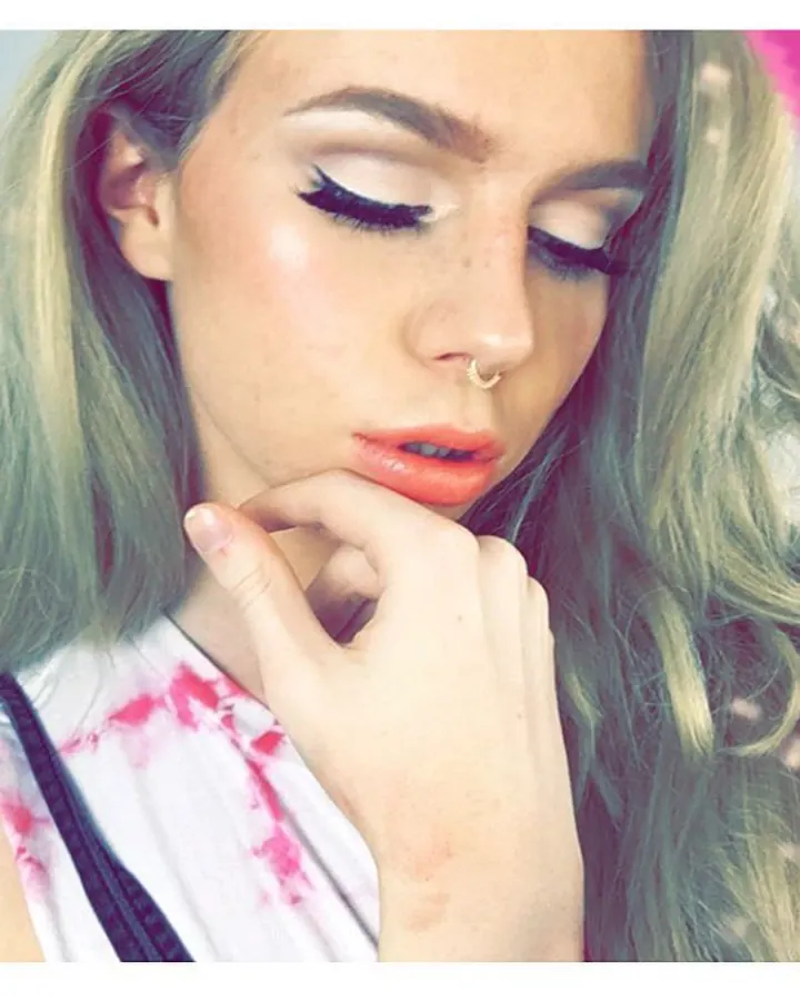 Femme aux cheveux blonds portant un haut blanc et noir, posant de profil avec un regard vers le côté, devant un fond naturel.