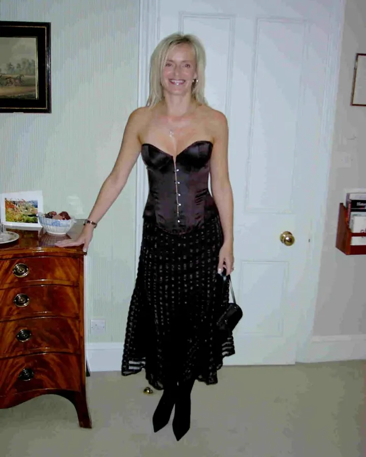 Femme aux cheveux blonds en corset noir et jupe longue, souriante, dans un intérieur élégant.